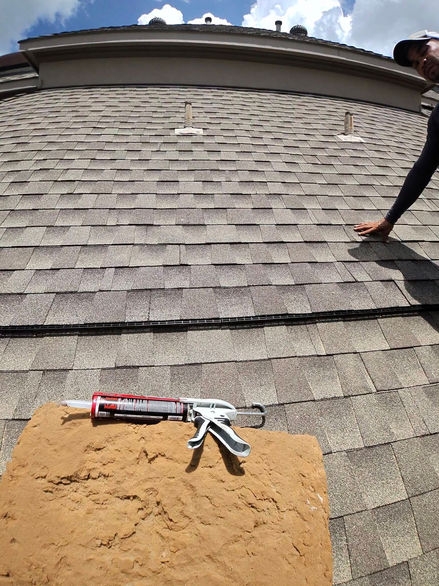 Roof shingles with a caulk gun and putty knife on a board, while someone works on the roof edge