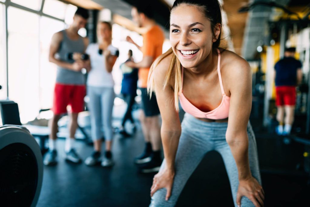 Smiling woman leaning forward in a gym, with people exercising in the background.
