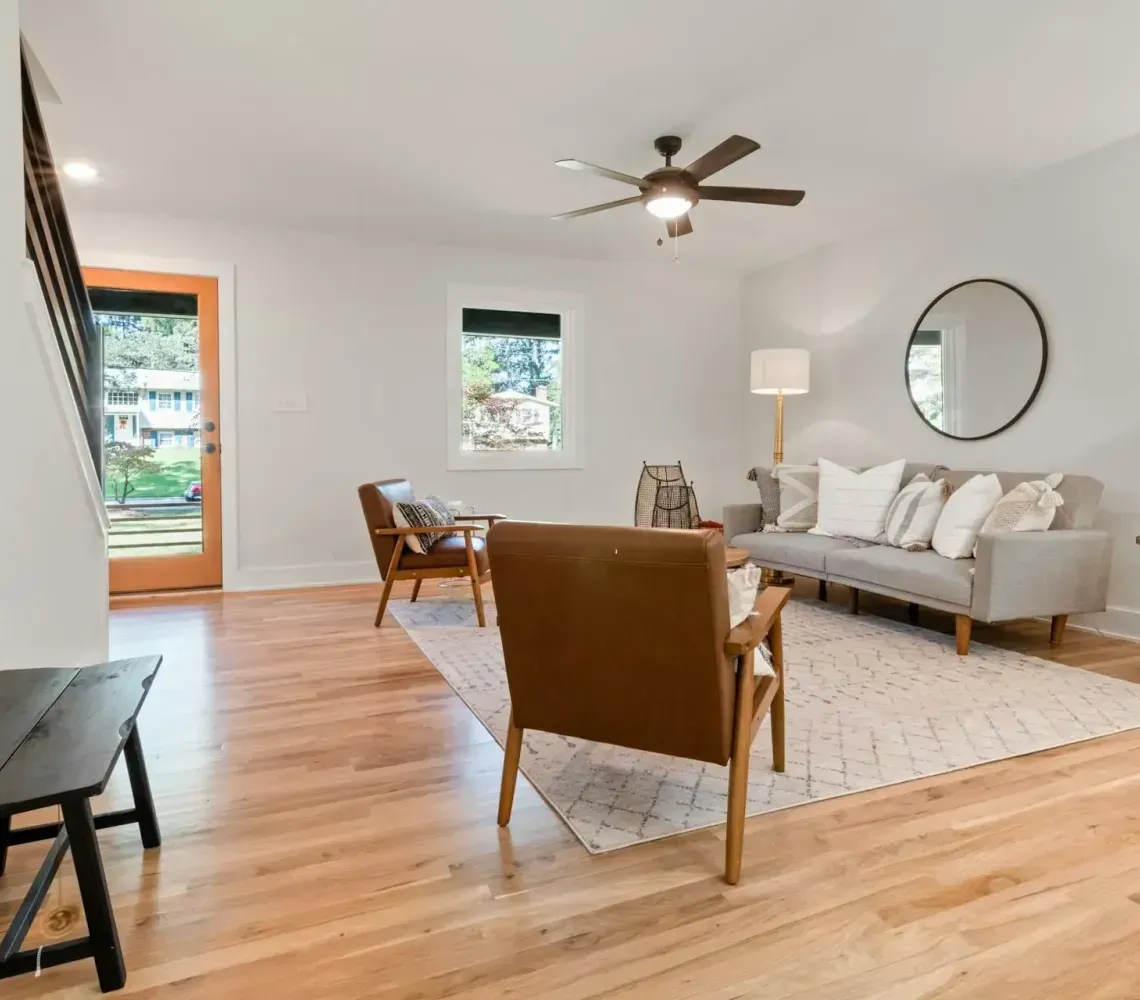 A bright living room with light wood floors, a gray sofa, brown leather accent chairs, a round mirror, and a wood door.