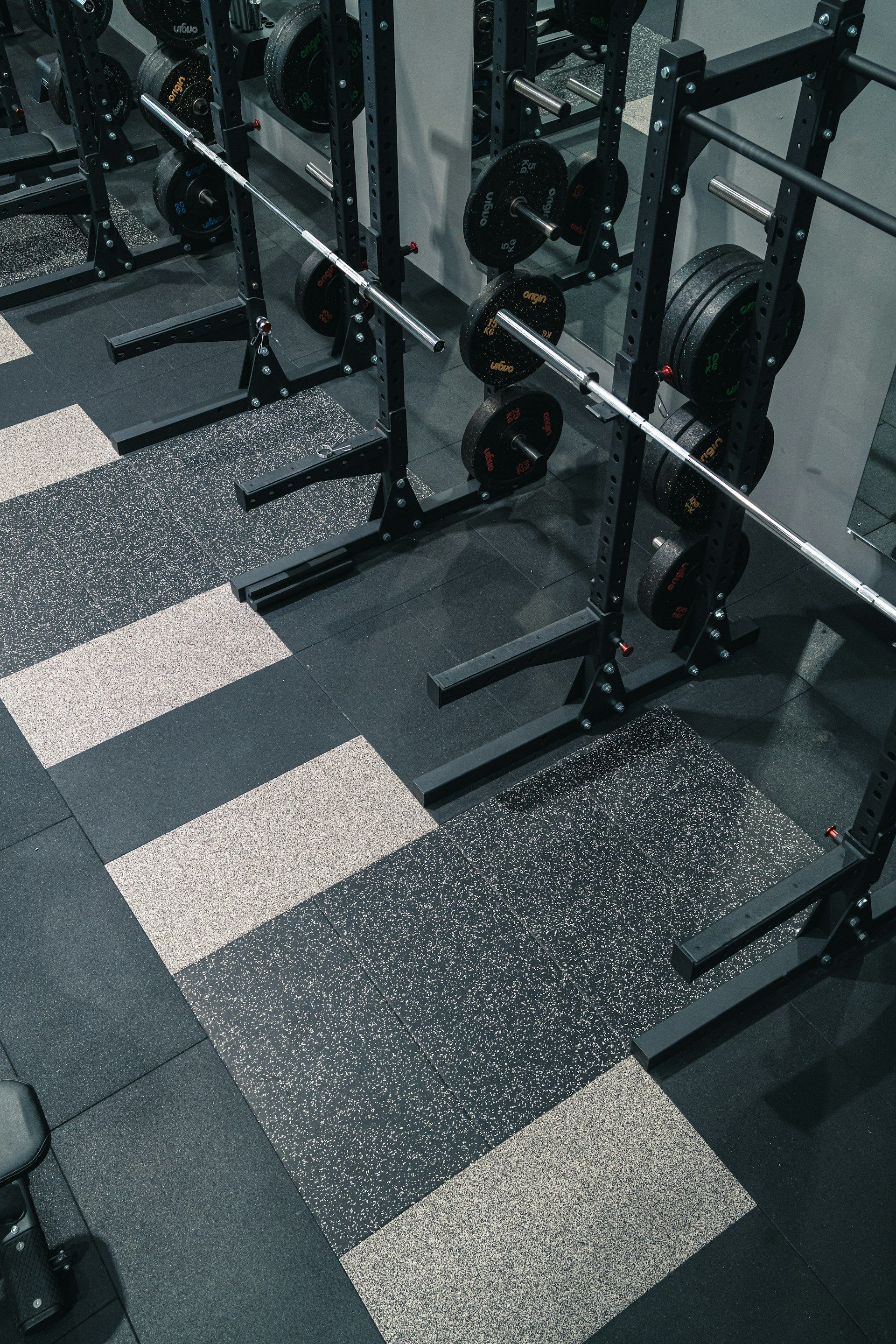 A row of empty black weightlifting racks with barbells in a gym with patterned, speckled rubber flooring.