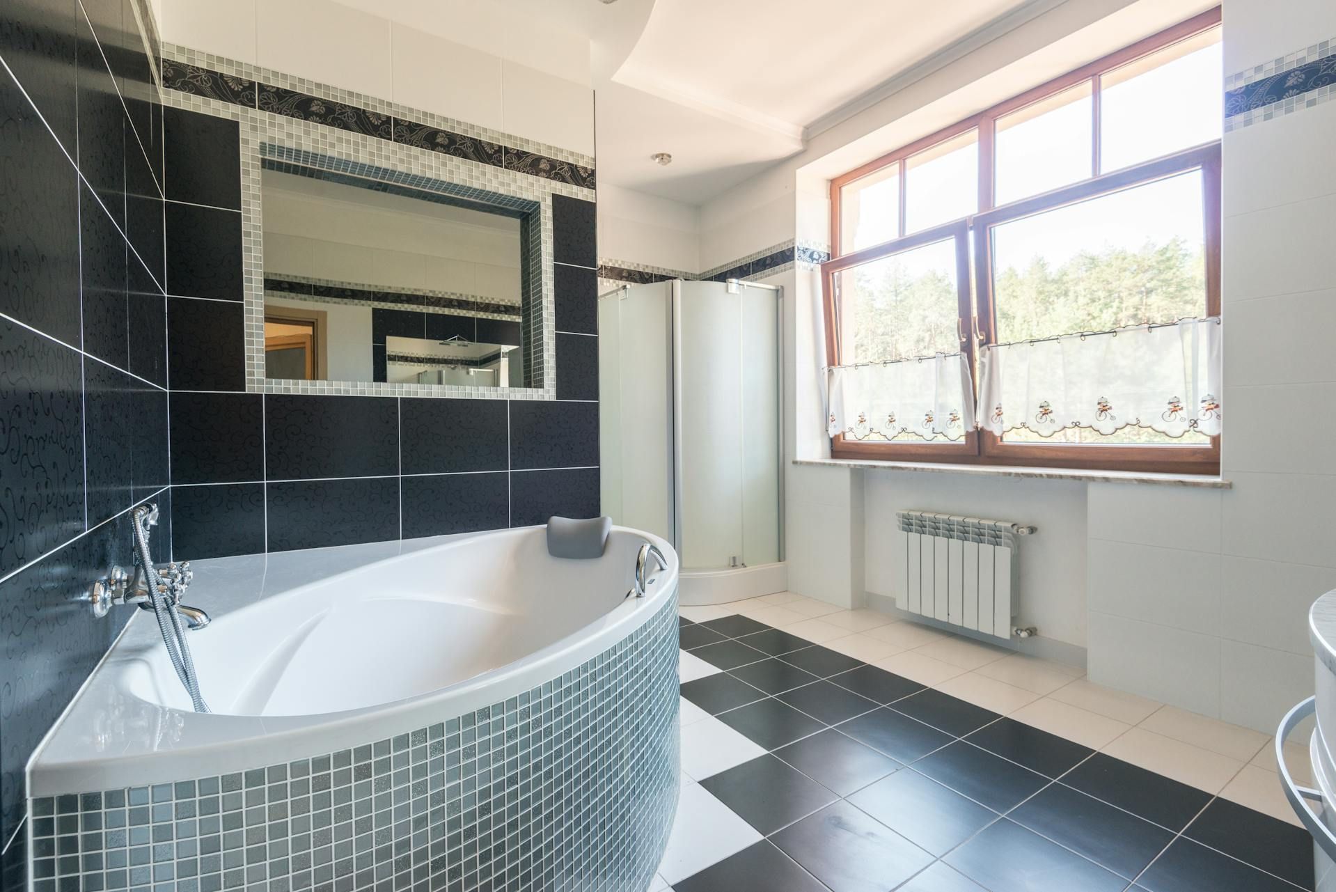 A modern bathroom featuring a white curved bathtub with mosaic tiling, a black-tiled wall, and a large window.