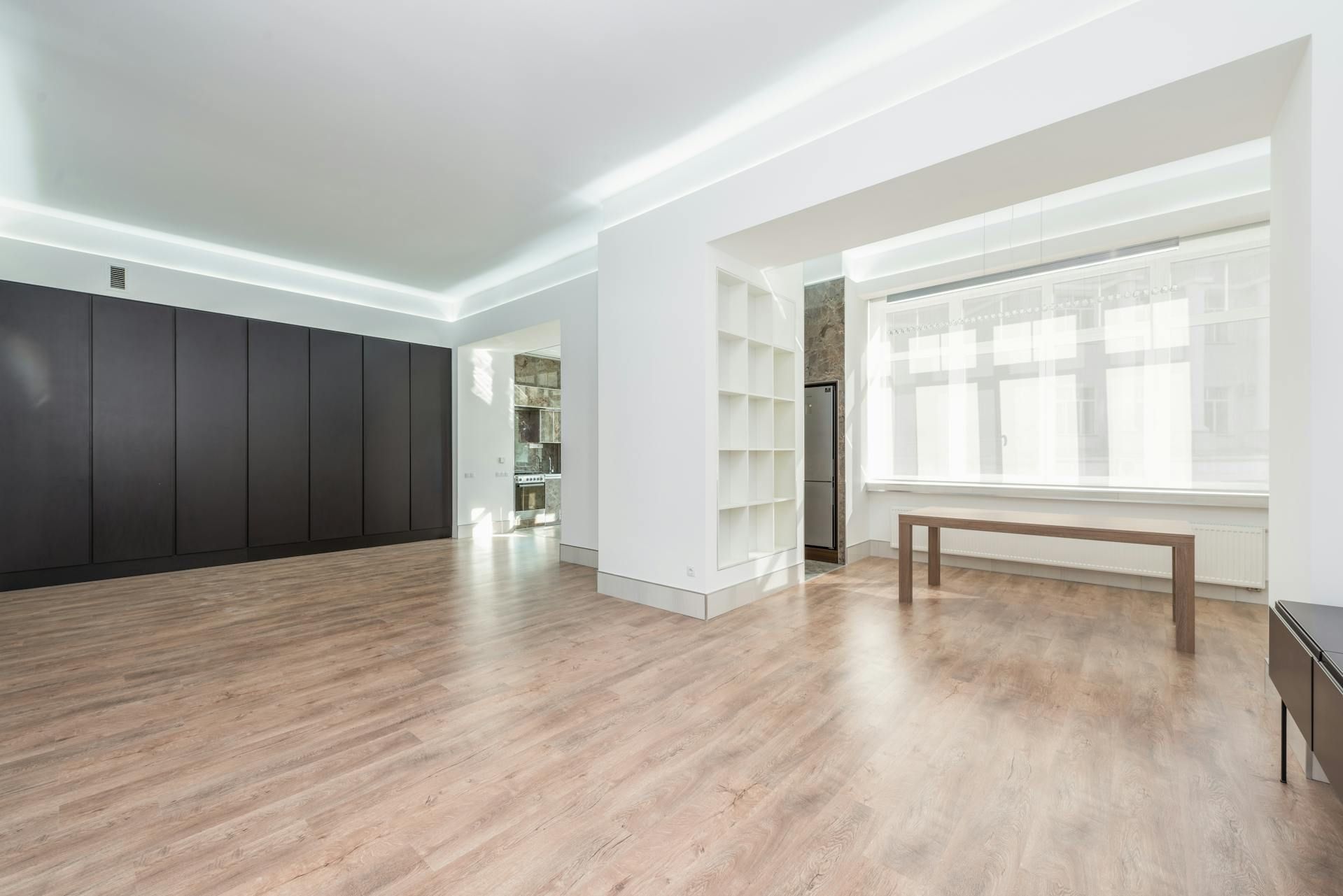 Bright empty living room with light wood floors, black cabinets, white shelves, and a bench by a large window.