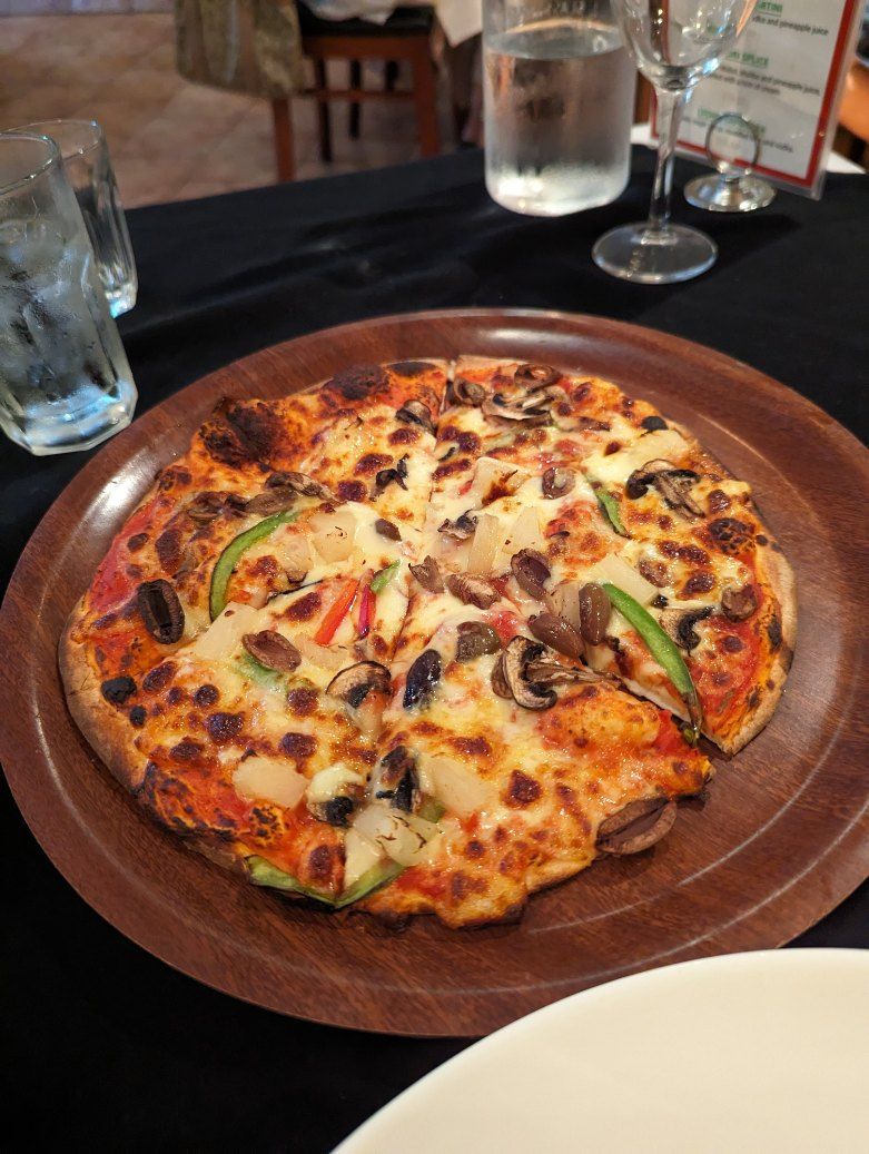 Cheesy pizza with vegetables on a wooden platter on a dark restaurant table, with drinks in the background — Romanas Italian Restaurant in Mermaid Beach, QLD