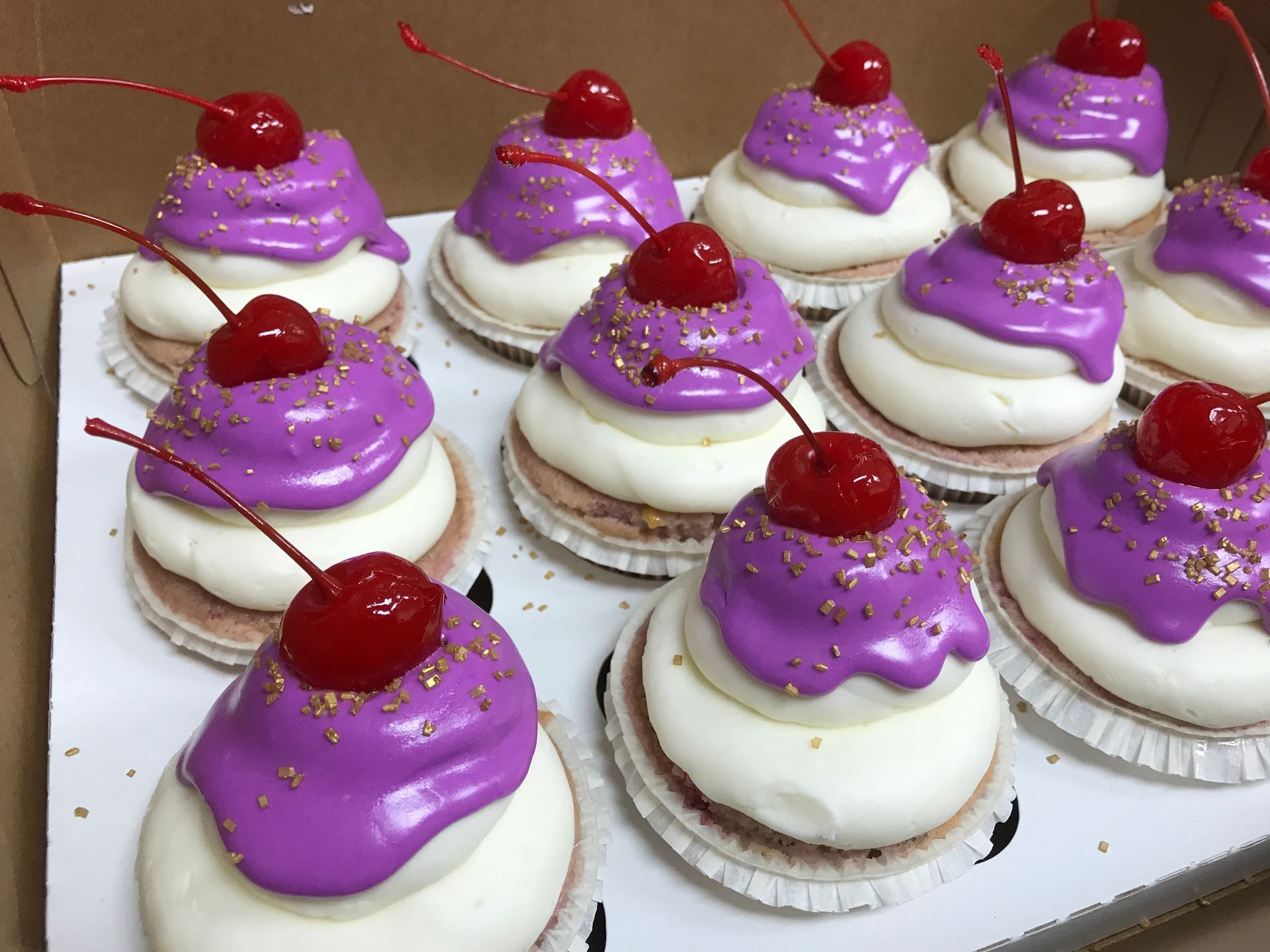 a box of cupcakes with purple frosting and cherries on top