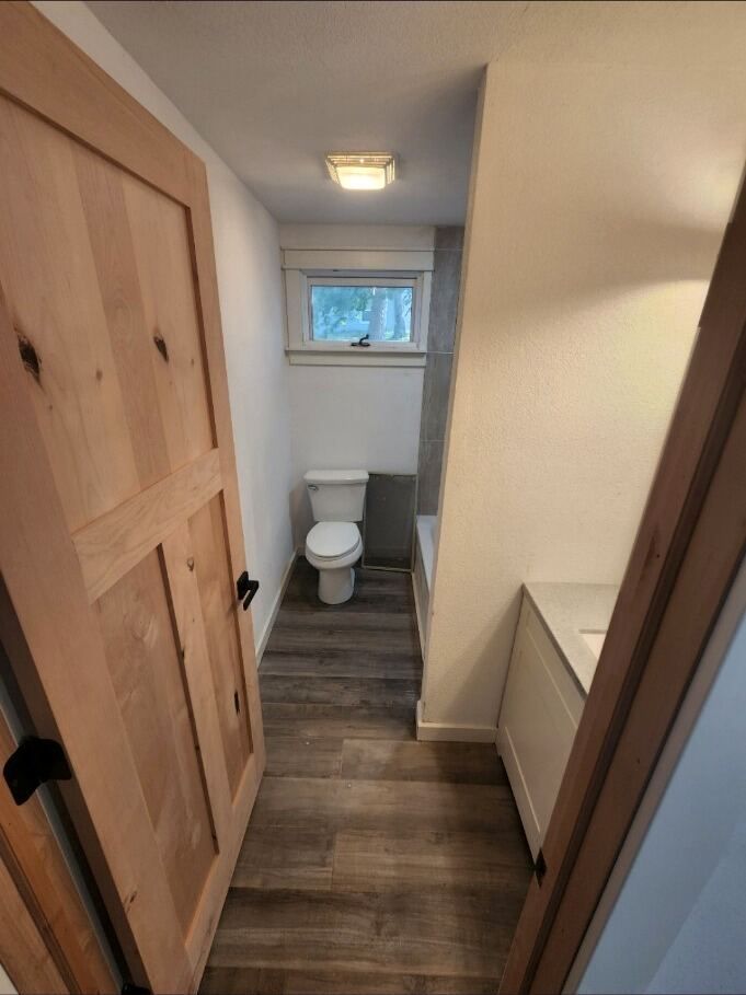 Narrow bathroom with toilet, wood-look floor, beige walls, and a small window at the end.