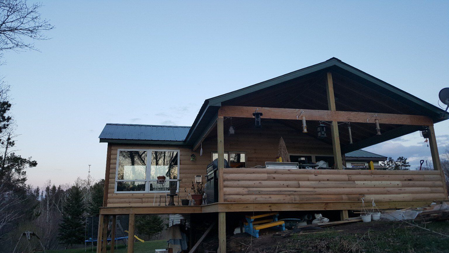 Wooden cabin with a large porch and blue roof at dusk, surrounded by trees.