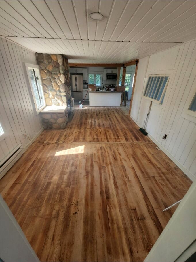 Empty room with wood-look floor, white paneled walls, stone fireplace, and a kitchen area in back.