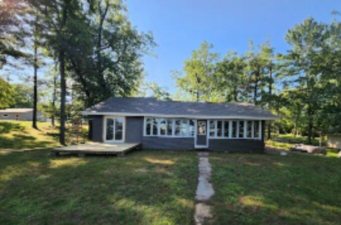 Small gray house with a front porch, centered stone path, and trees in a sunny yard.