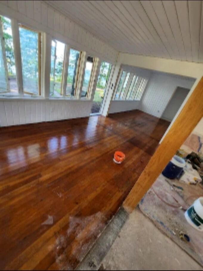 Empty sunroom with glossy wooden floor, large windows, and a small orange bucket near the center.