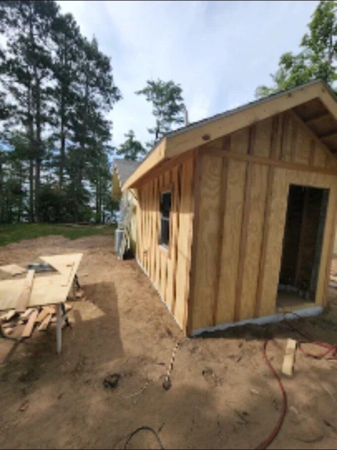 Wooden cabin under construction in a forested area, with tools and dirt ground nearby.