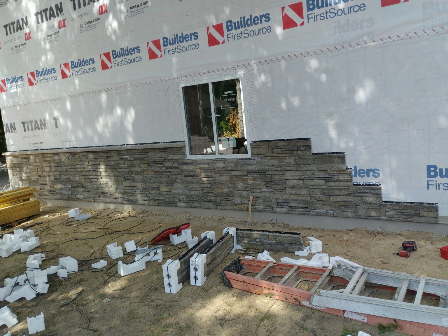 Construction materials and ladder laid out beside a building under renovation with wrapped walls and stone base.