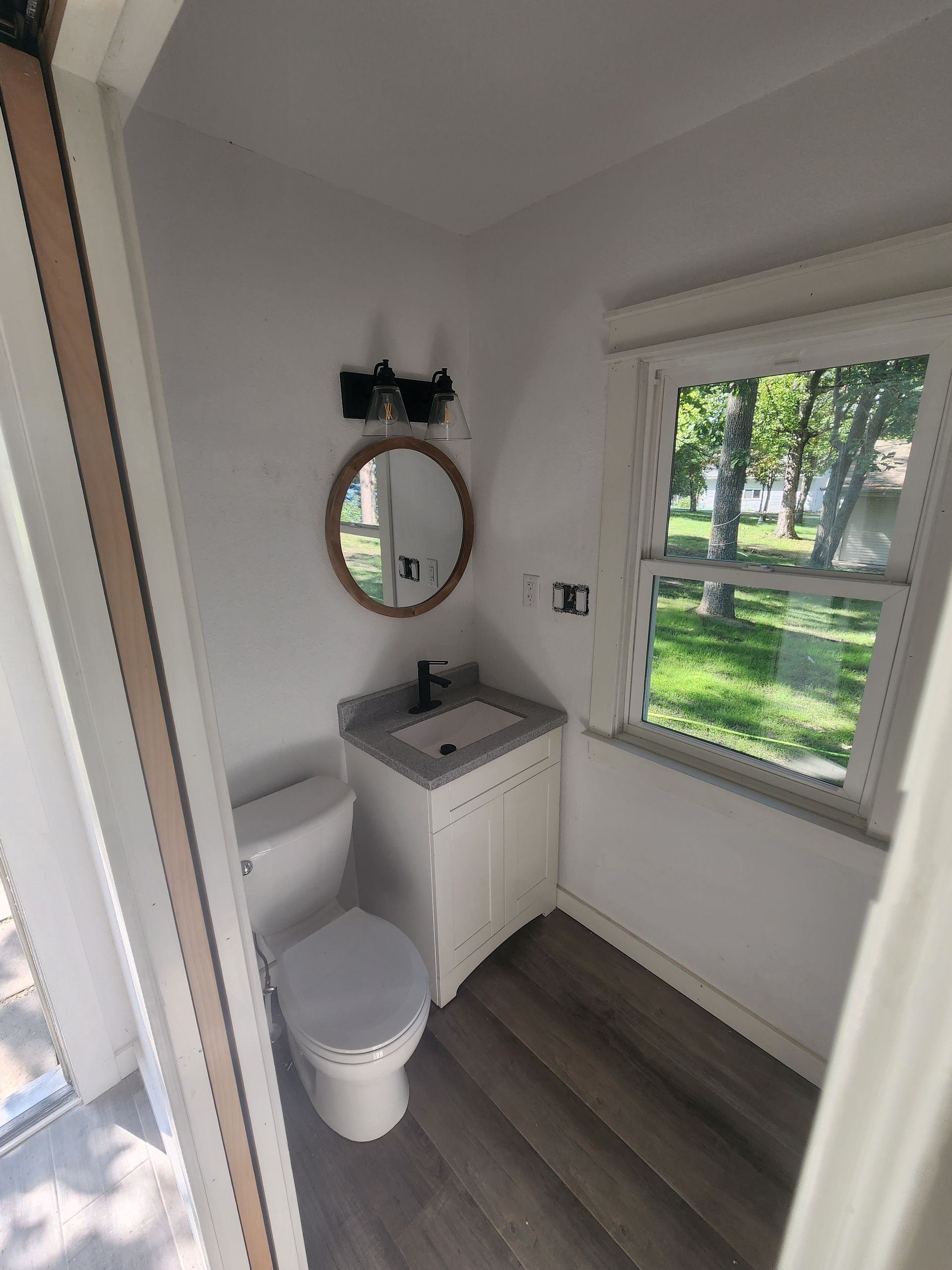 Small bathroom with toilet, sink, round mirror, and window overlooking greenery.