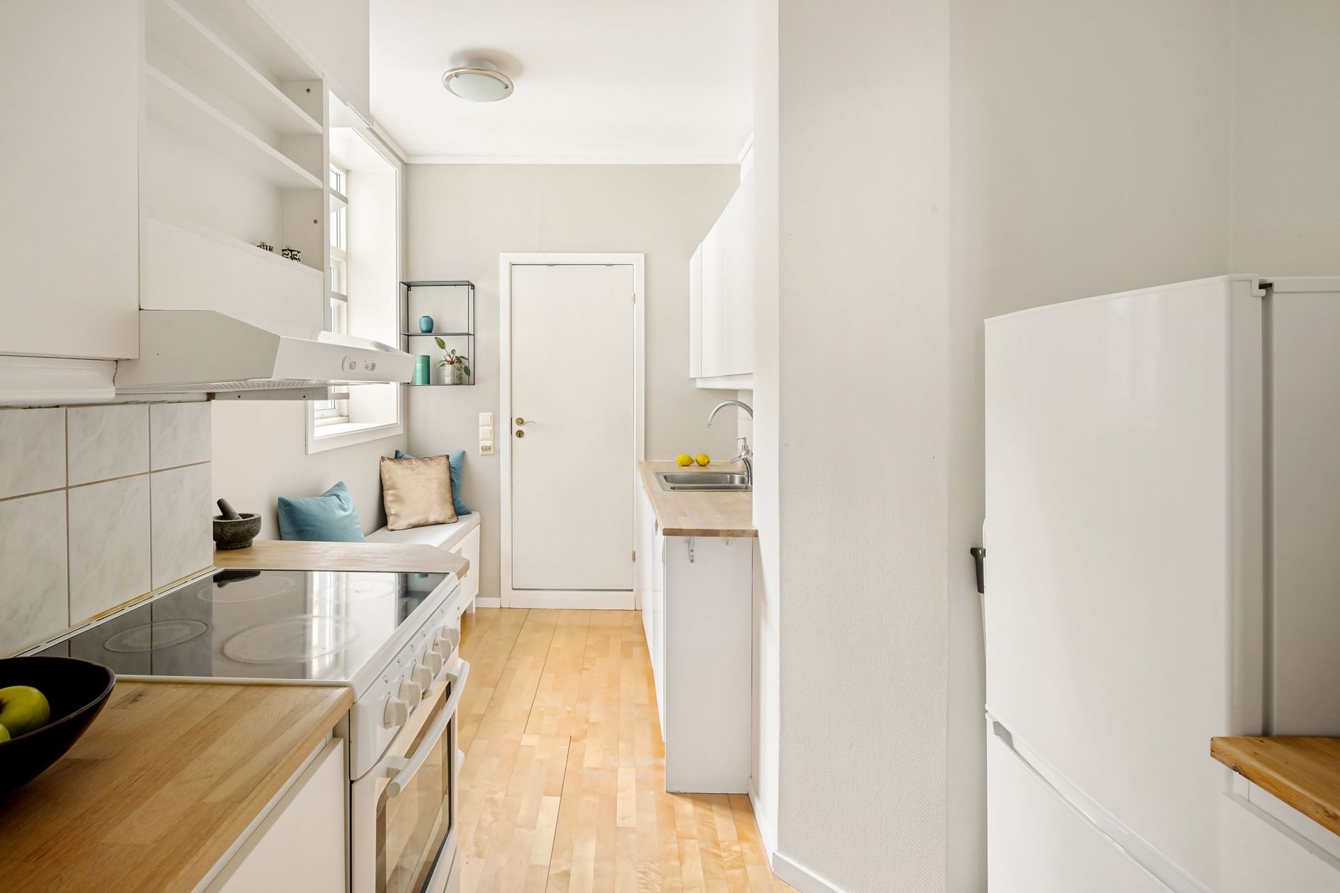 Narrow white kitchen with wooden floor, sink and counter on the left, refrigerator on the right.