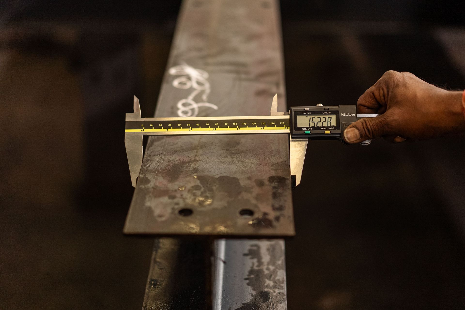 Hand measuring a metal beam with a digital caliper and yellow tape measure