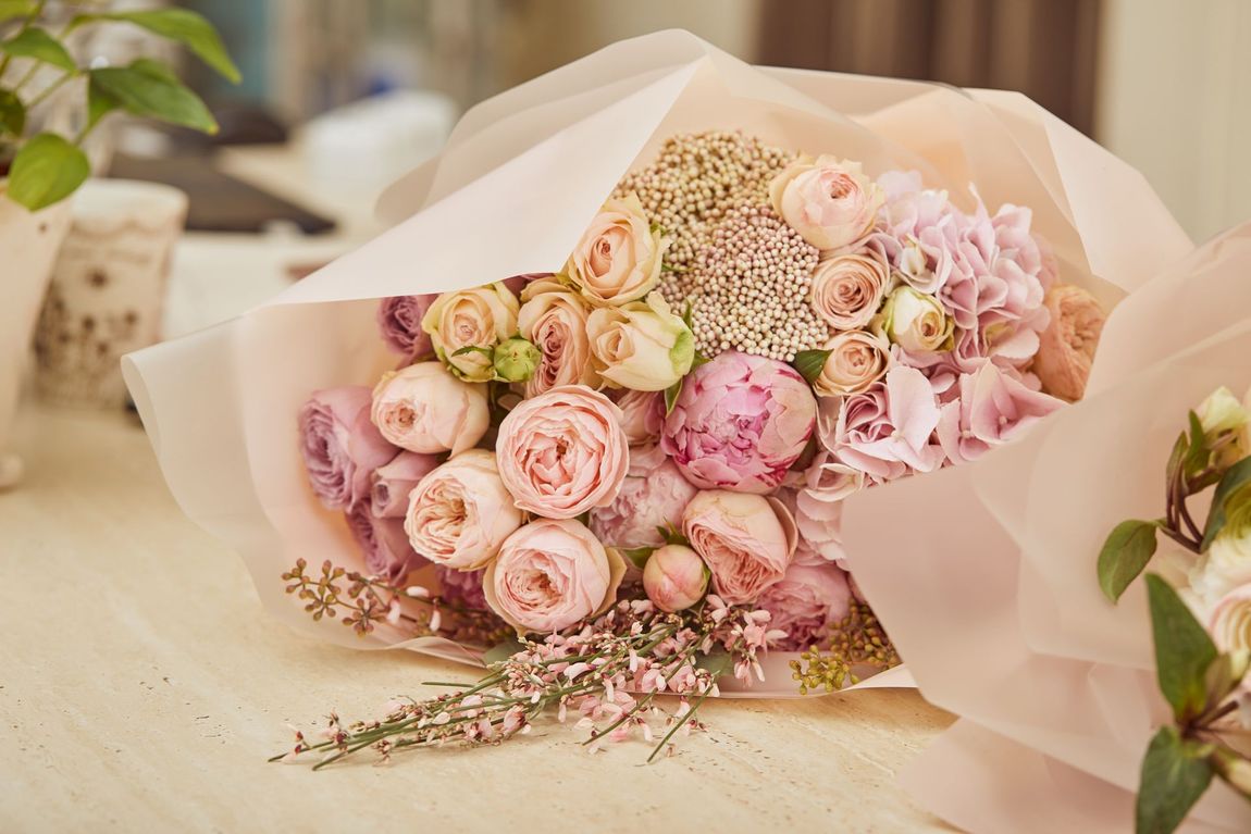 Un bouquet di rose e ortensie rosa tenue e pastello, avvolto in una delicata carta rosa chiaro su uno sfondo bianco.