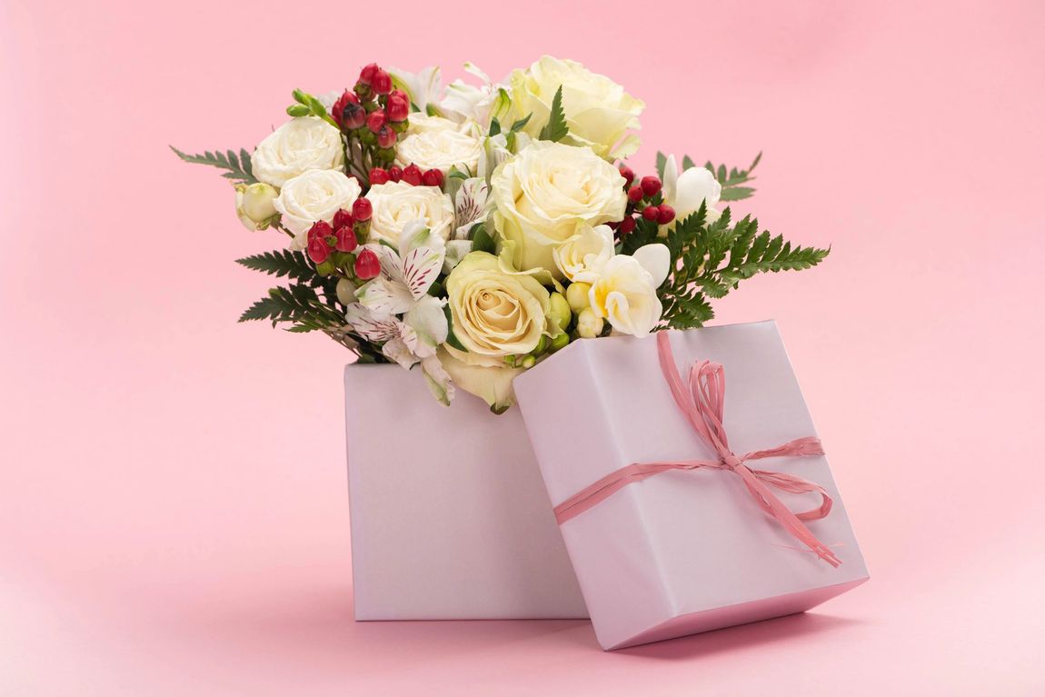 Un bouquet di rose bianche e bacche rosse in una confezione regalo lilla chiaro con un nastro, su uno sfondo rosa.
