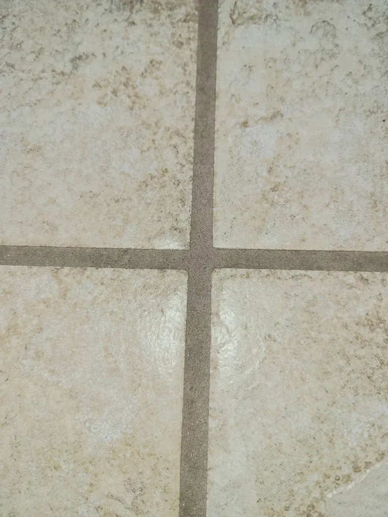 Close-up of beige, textured ceramic floor tiles separated by dark brown grout lines in a grid pattern.