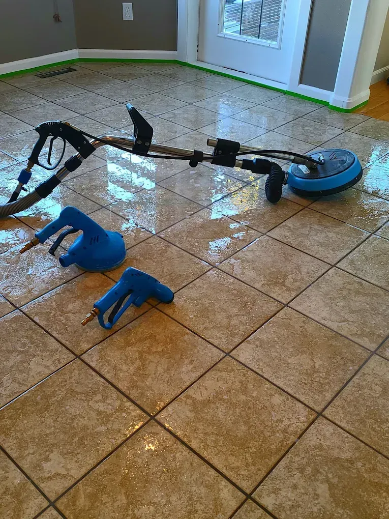 Tile floor cleaning tools, including a rotary surface cleaner and two blue spray guns, on a wet, tiled residential floor.
