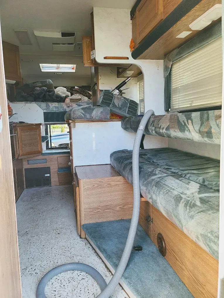 Interior of a camper featuring a patterned bench seat, wooden cabinetry, and a vacuum hose extending across the floor.
