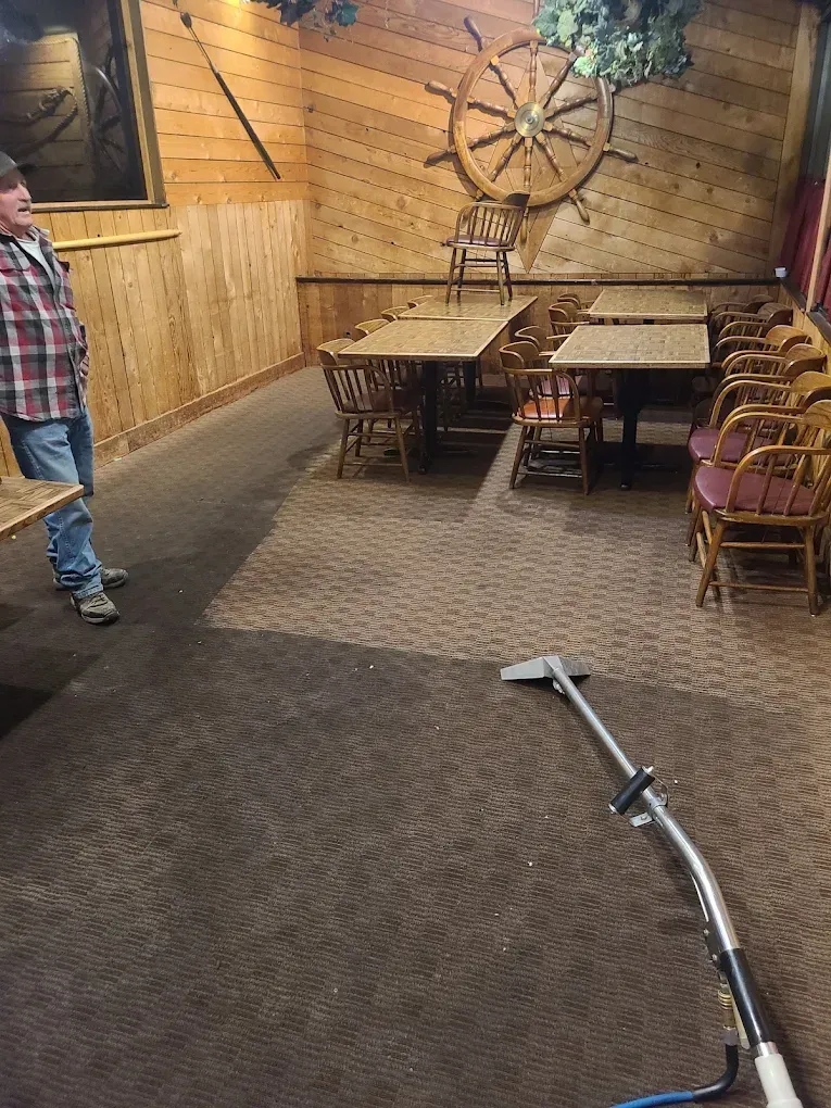 A person stands in a restaurant with wood-paneled walls and a ship’s wheel, as a carpet cleaning wand rests on the floor.
