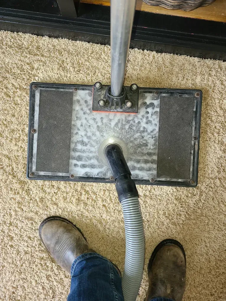 A top-down view of a carpet cleaning wand on beige carpet, with a person's work boots visible in the foreground.