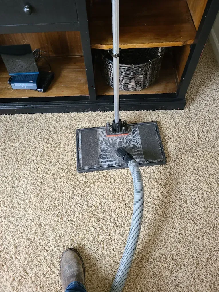 A person cleans a beige carpet using a long-handled floor cleaning tool connected to a vacuum hose.