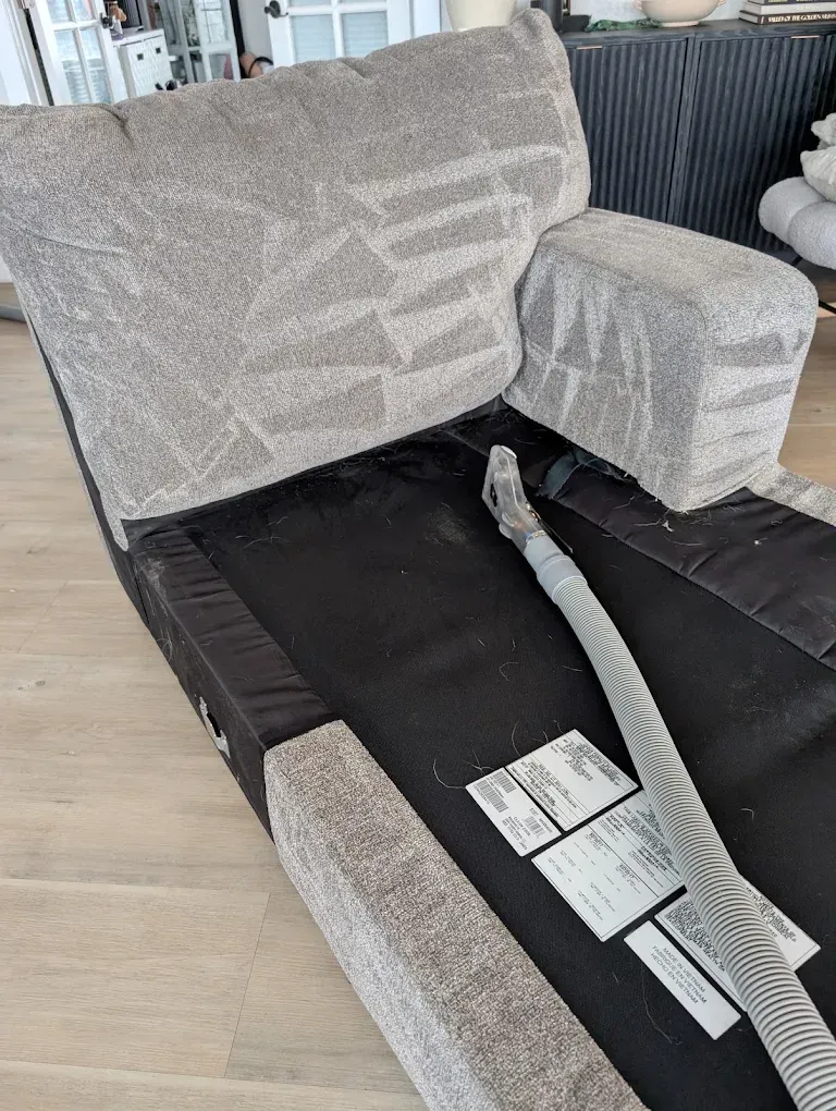 A vacuum cleaner hose rests on the exposed black seat base of a light-colored, textured fabric armchair.