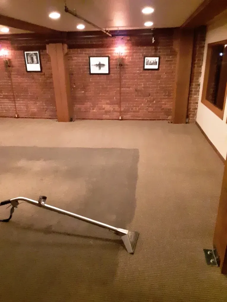 A carpet cleaning wand rests on a partially cleaned beige carpet in a room with exposed brick walls and framed artwork.