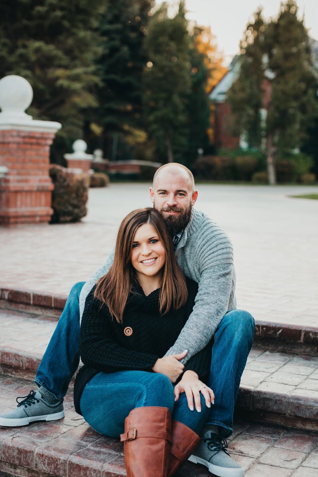 A couple sits on brick steps, smiling at the camera, wearing sweaters and jeans outdoors near a brick building.