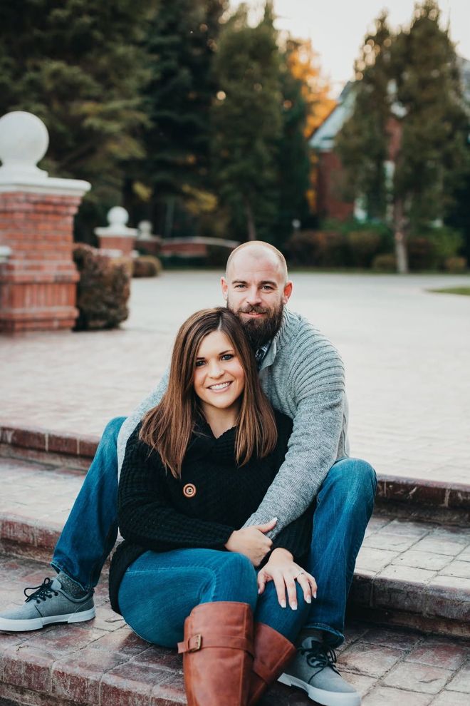 A couple sits on brick steps, smiling at the camera, wearing sweaters and jeans outdoors near a brick building.