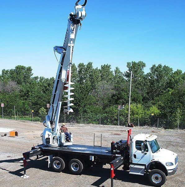 A white utility truck with a mounted derrick digger and auger extended upward in an outdoor gravel lot.
