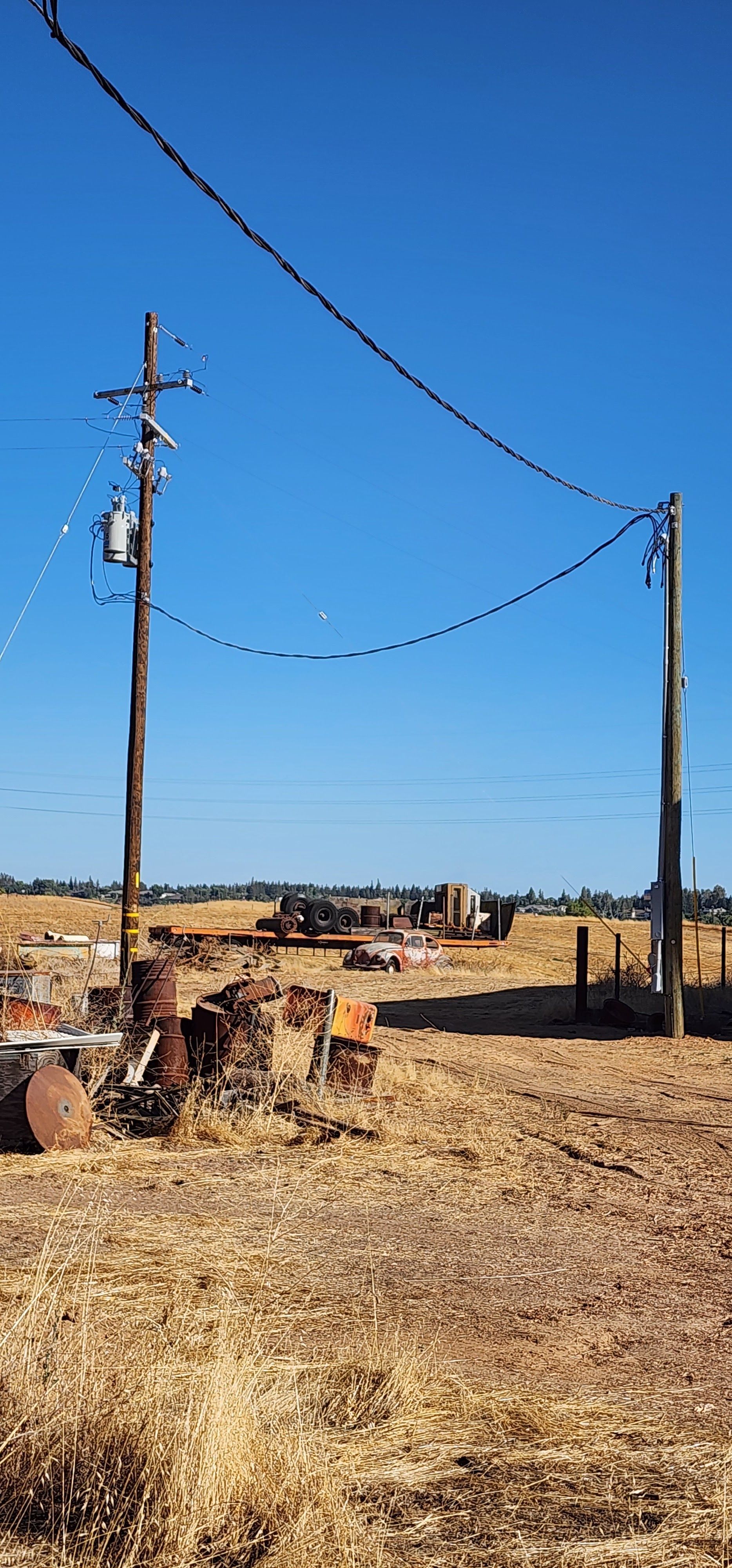 Two wooden utility poles connected by a line stand in a dry, harvested field under a clear blue sky.