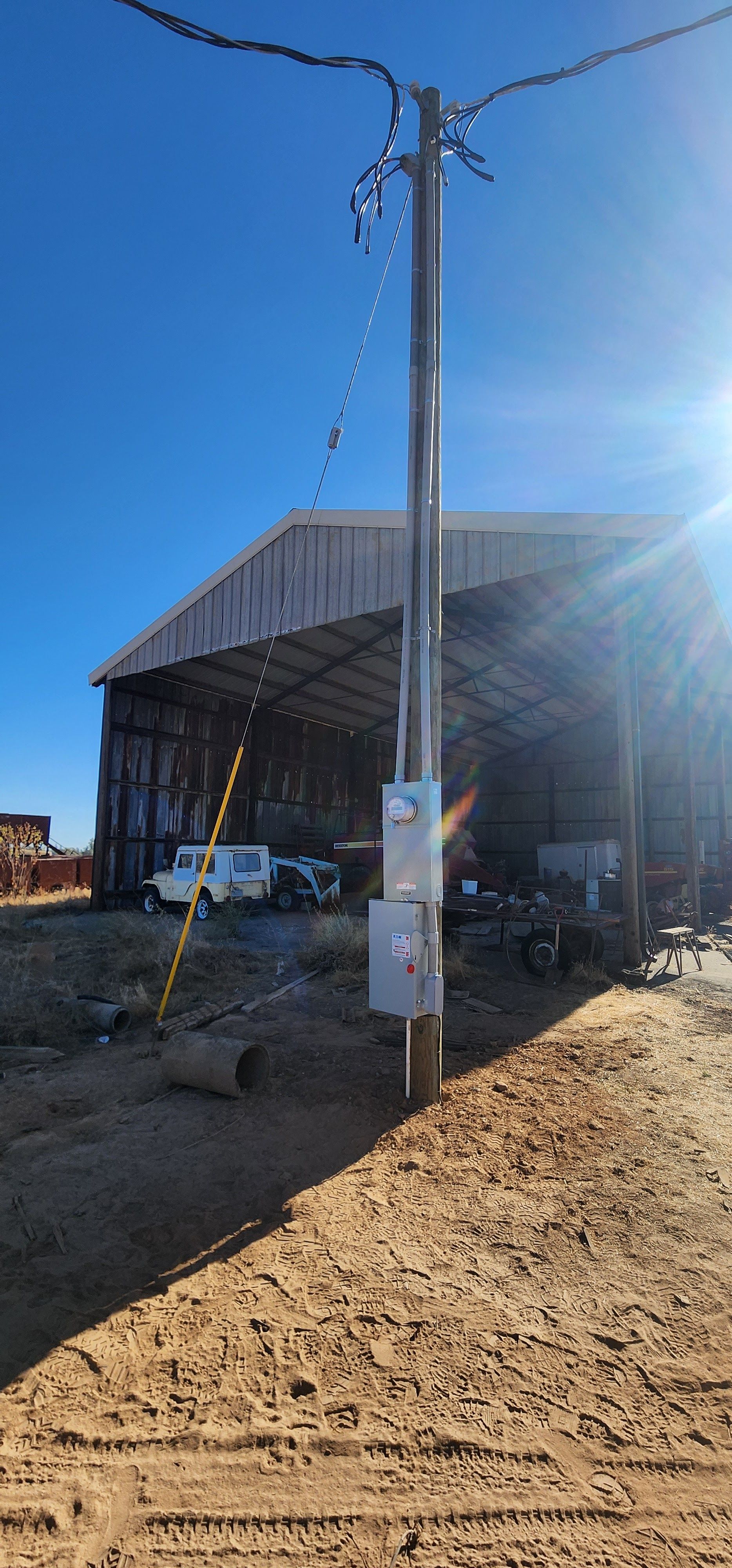 A wooden utility pole with two electrical boxes stands in a dirt lot in front of a large barn structure under a blue sky.