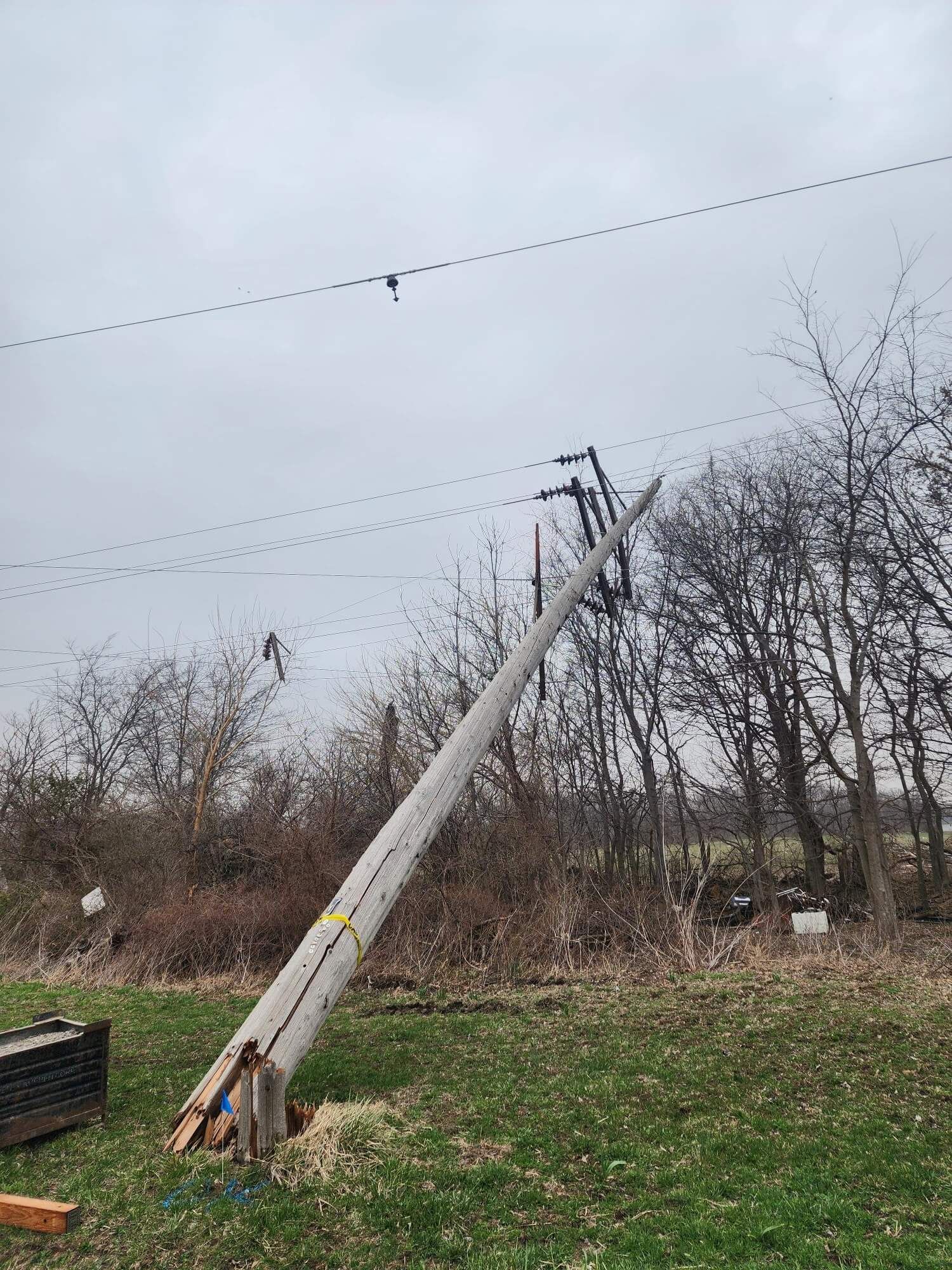 A utility pole leans at a sharp angle over grass, supported by electrical lines against a cloudy sky.