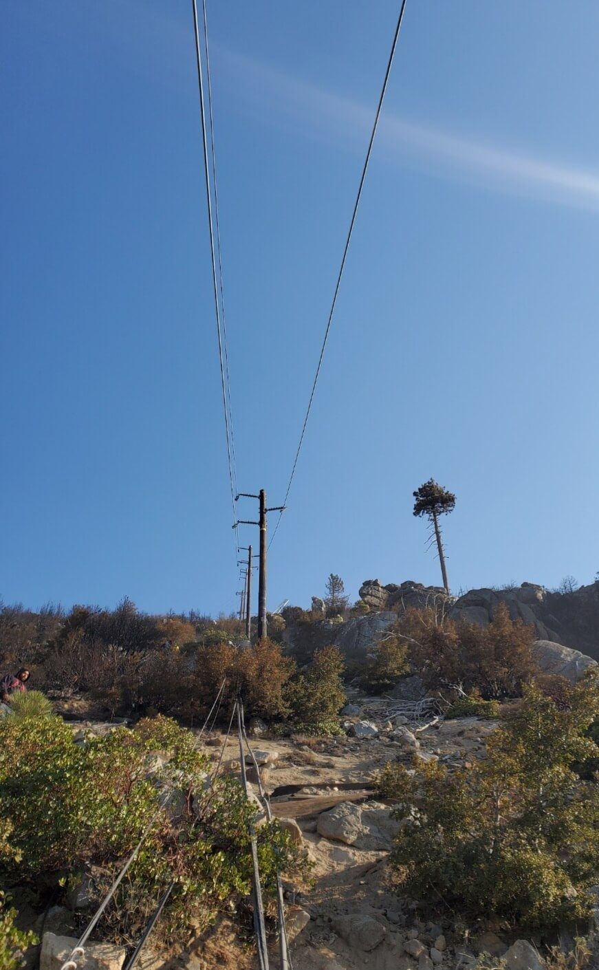 Utility poles and power lines run up a rocky, sunlit hillside against a clear blue sky.