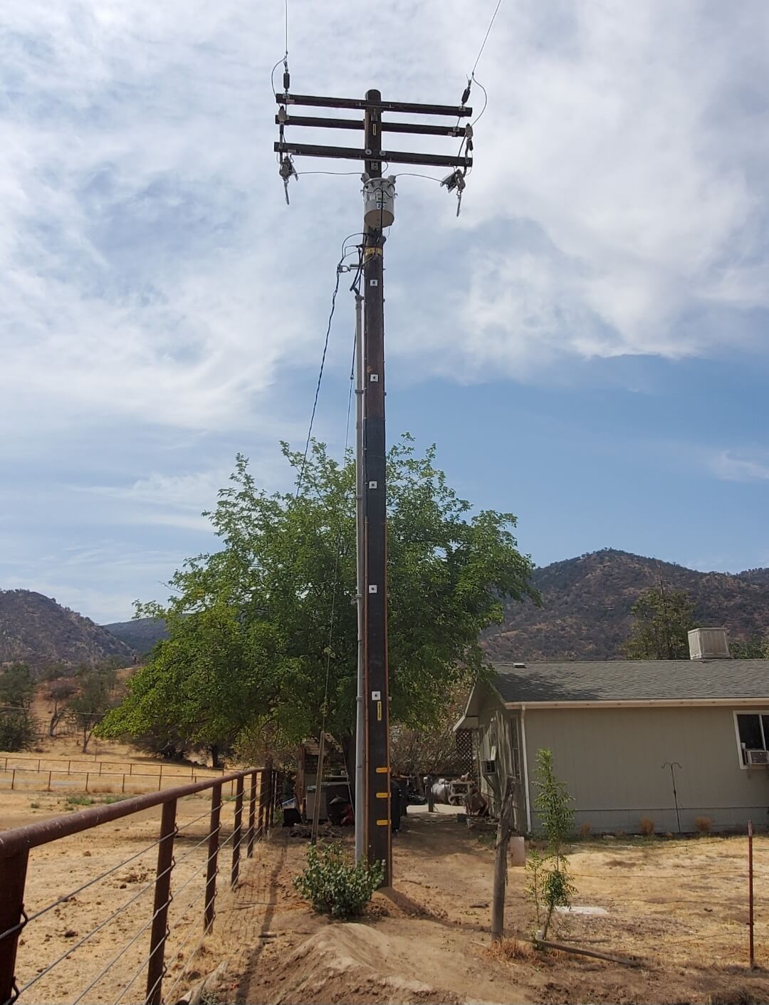 A wooden utility pole with a transformer stands in a dry, mountainous landscape next to a house and a fenced yard.
