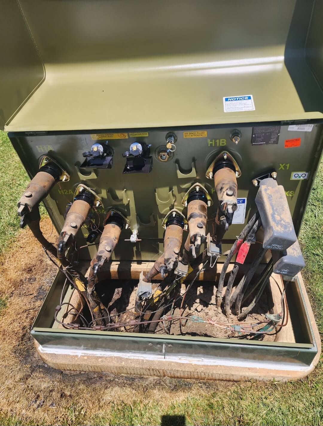 An open green pad-mounted electrical transformer with multiple heavy-duty power cables connected to its internal terminals.