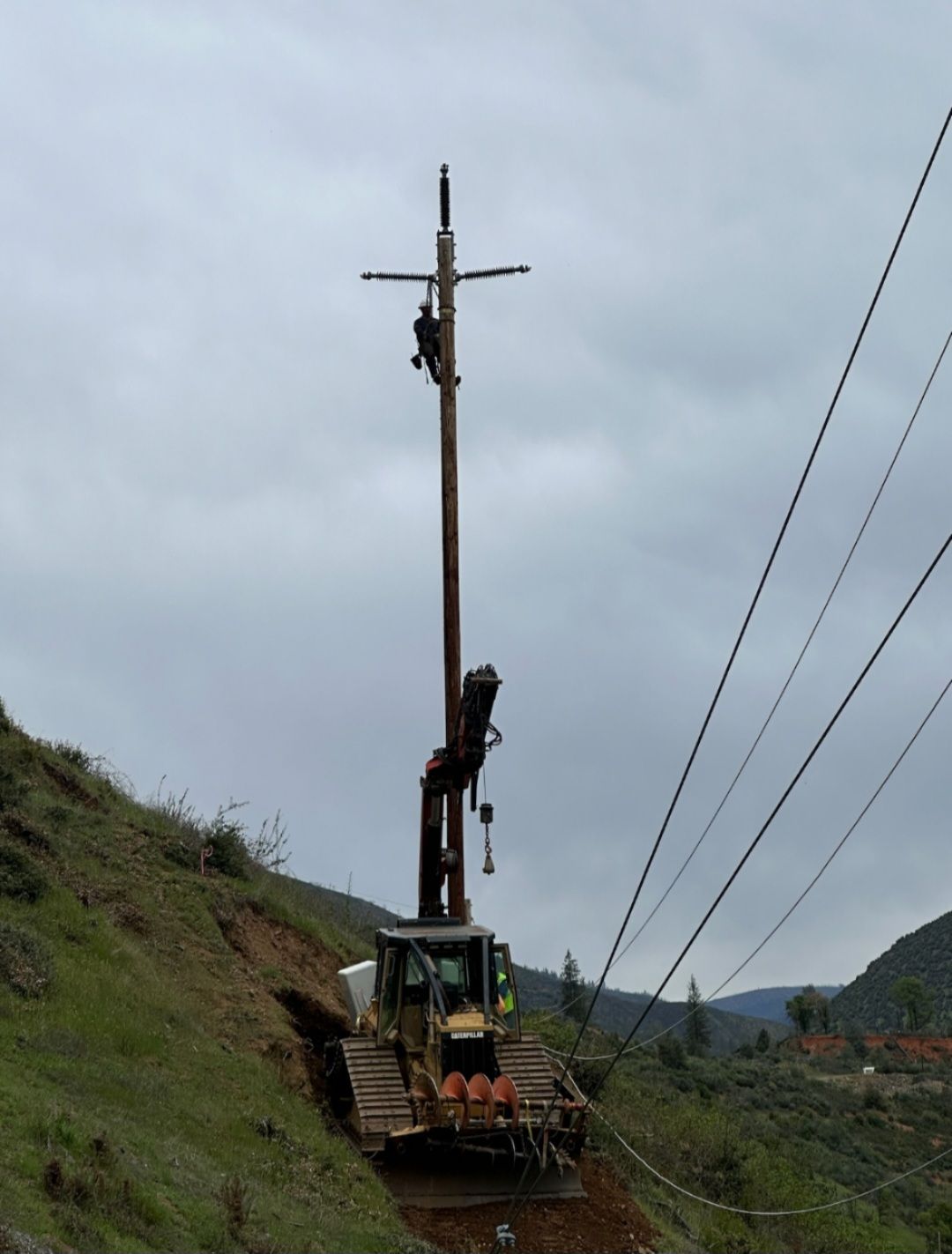 A worker stands on a tall utility pole being held vertically by a tracked crane on a hillside under a cloudy sky.