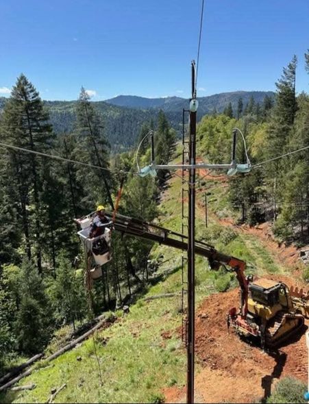 A utility worker in a bucket truck lift performs maintenance on power lines near a utility pole in a mountainous forest.