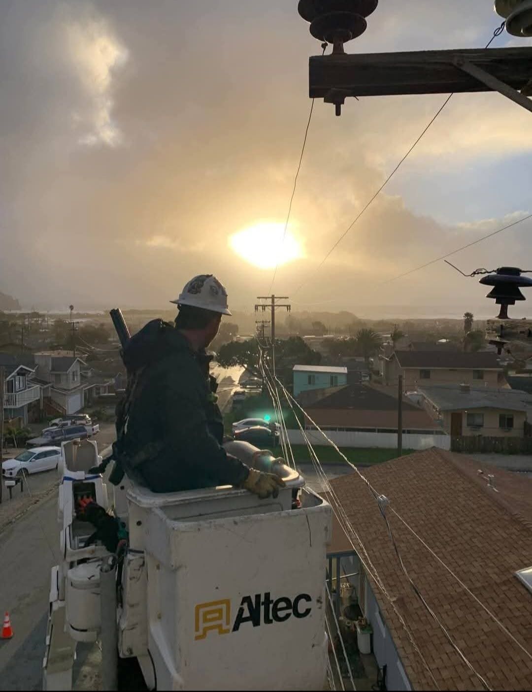 A utility worker in a bucket truck works on power lines at sunset above a residential neighborhood.