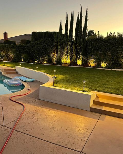 Illuminated backyard patio with pool, curved wall, lit lawn, and tall cypress trees at dusk