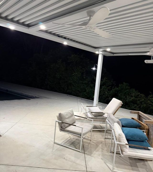 Outdoor patio at night with white lounge chairs, a table, and a ceiling fan under a covered roof.