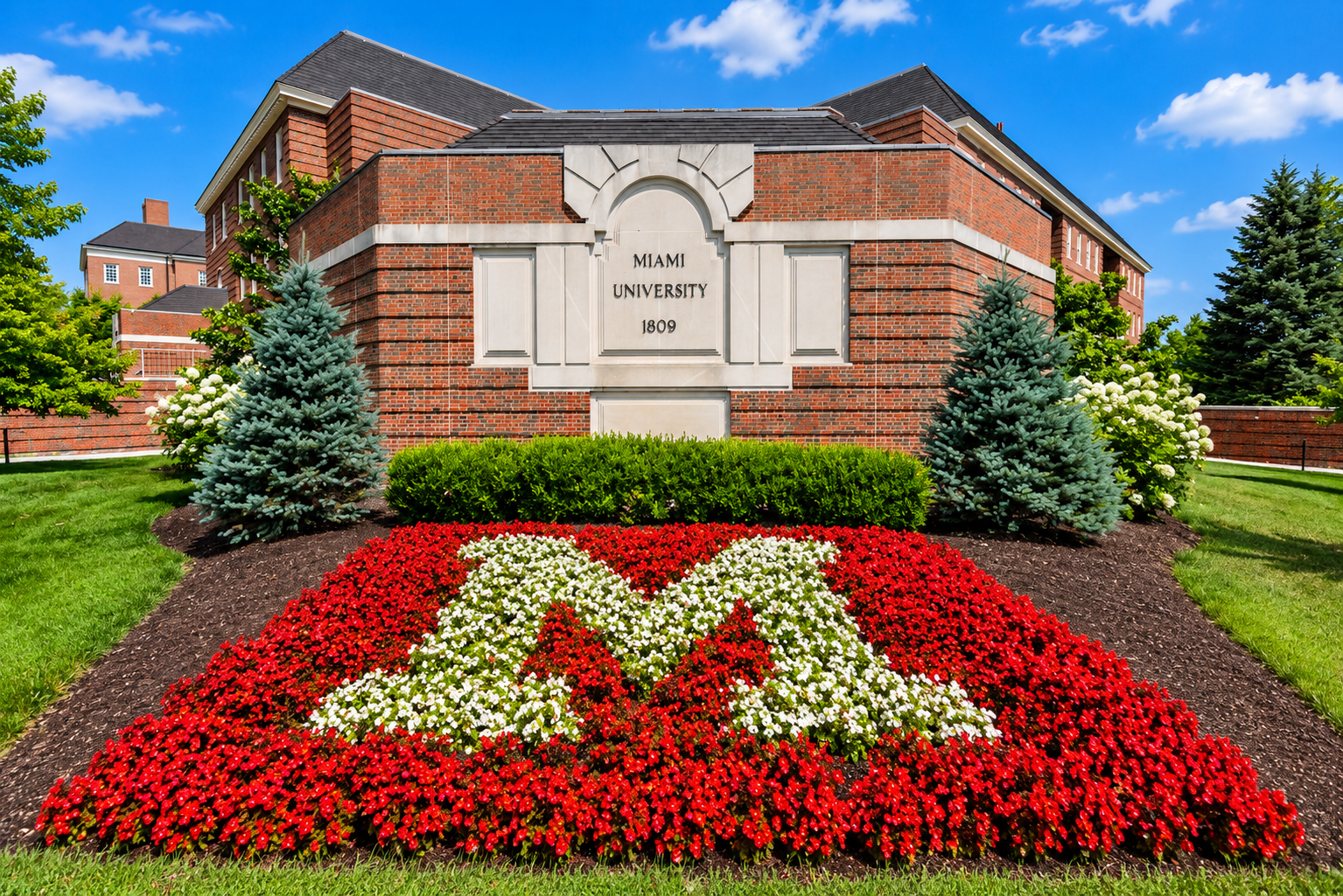 Brick building with a white M plaque and a red flowerbed in front, framed by evergreen shrubs