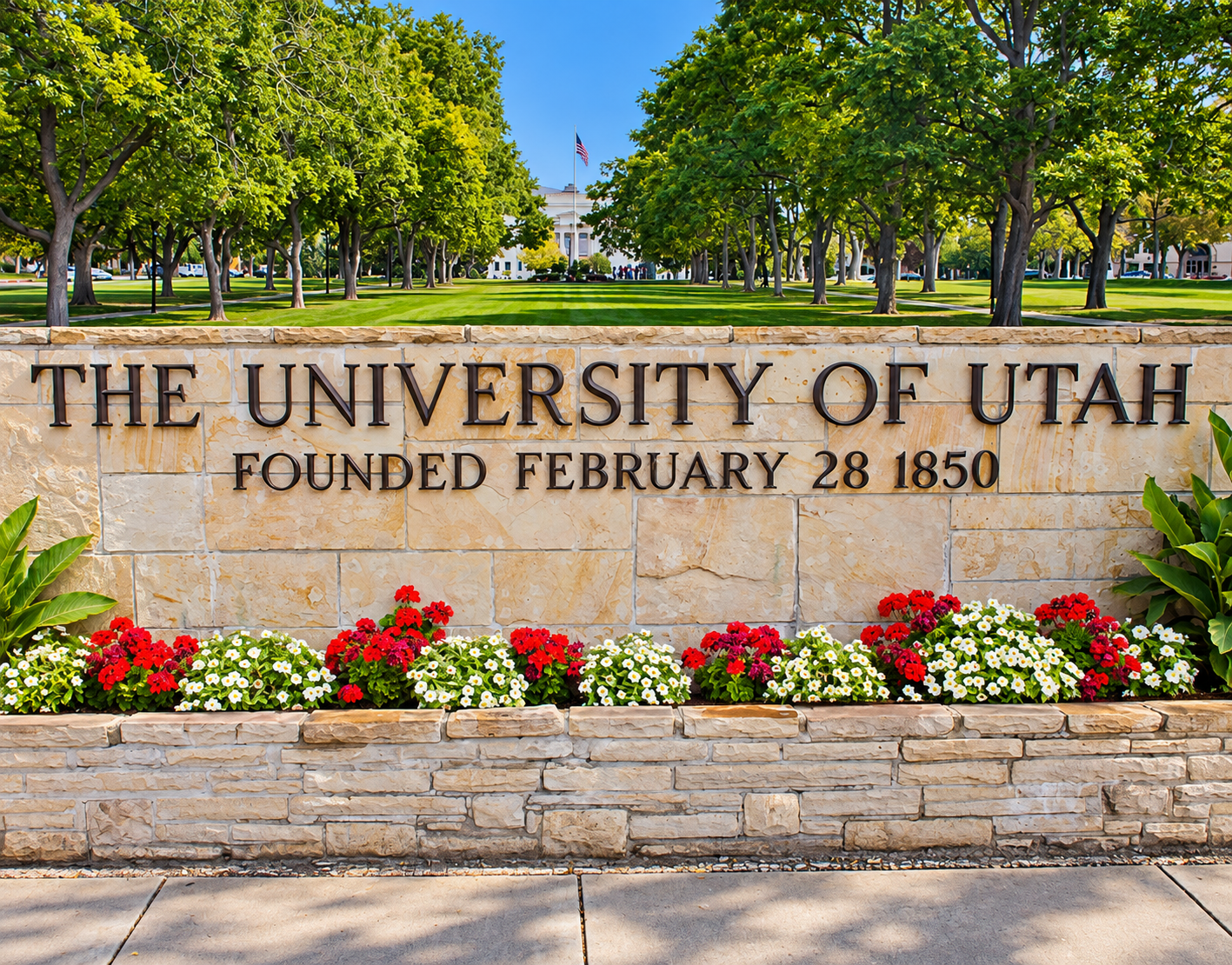 The University of Utah stone sign with flowers, trees, and a campus walkway under a bright blue sky