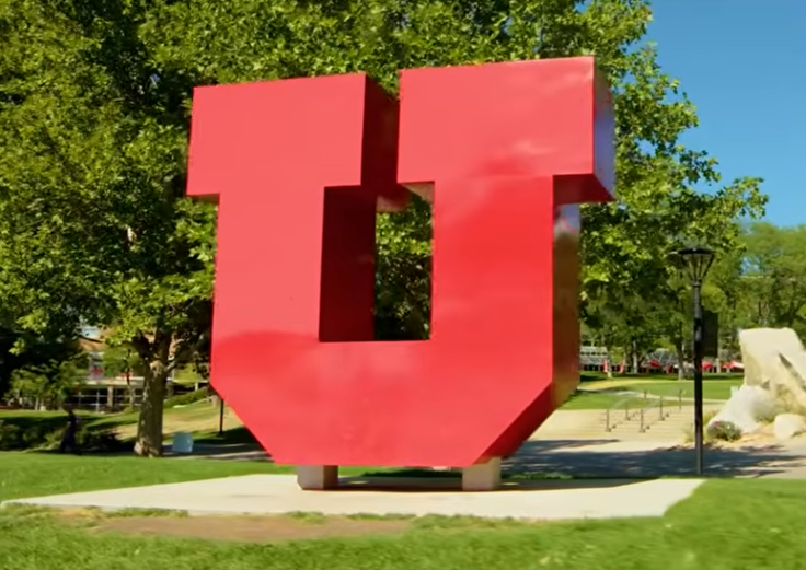 Red University of Utah “U” sculpture on a campus lawn with trees in the background