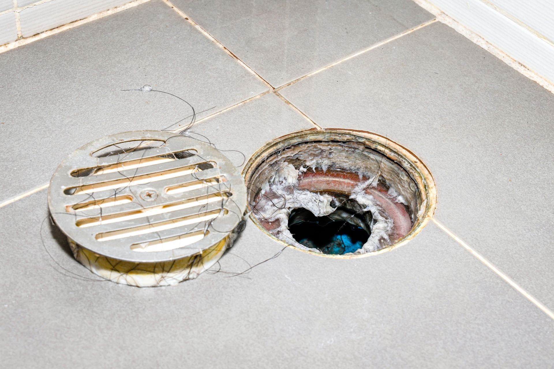 Two open floor drain covers on gray tile, one rusty and broken, one metal grate lifted aside