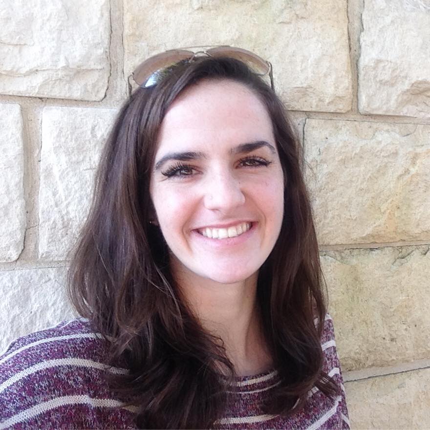 A woman wearing sunglasses is smiling in front of a brick wall.