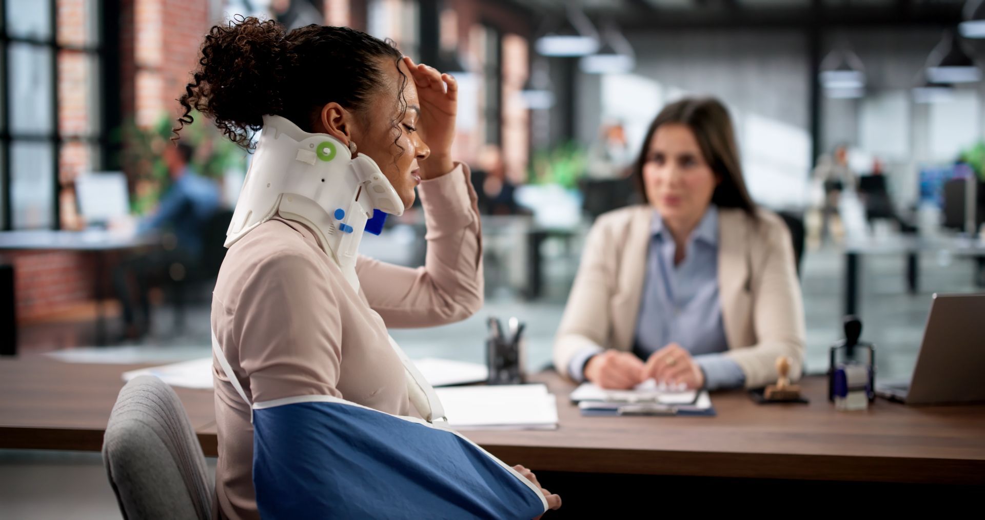 Person with neck brace and blue sling sitting in office, touching head while another person consults across table Person with neck brace and blue sling sitting in office, touching head while another person consults across table