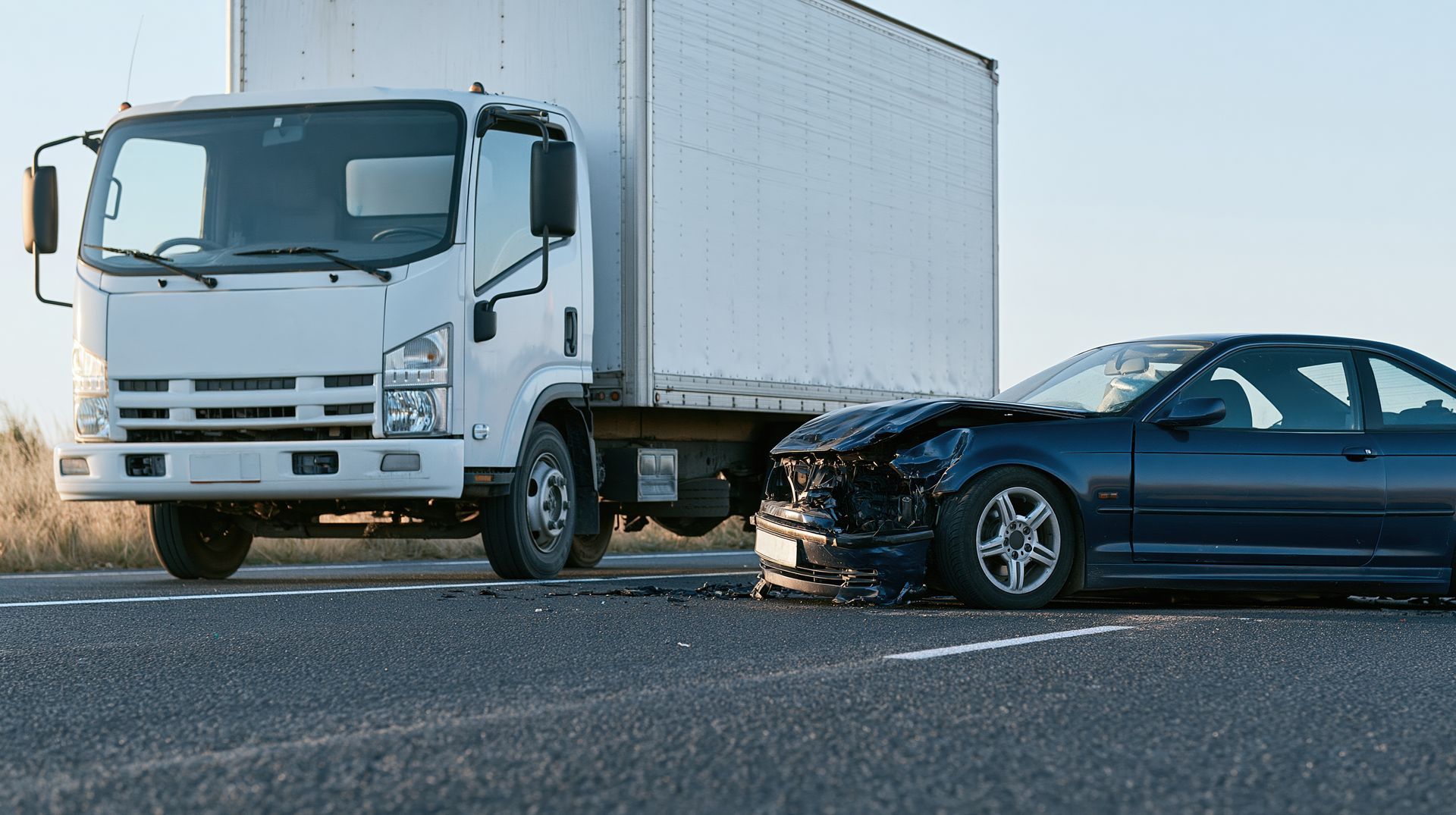 White box truck and dark sedan collide on a road, with the car’s front badly damaged.