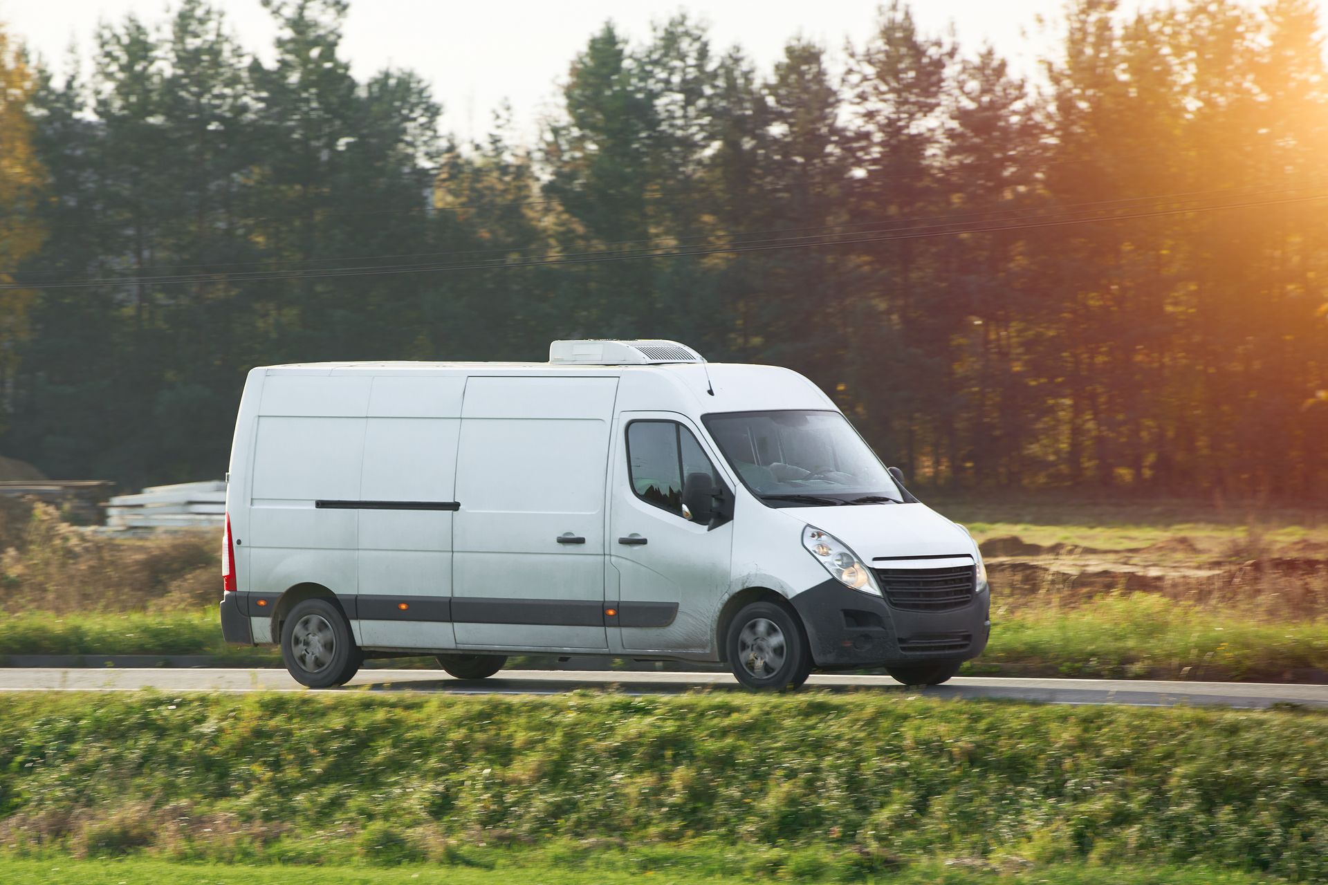 White cargo van driving on a rural road beside a grassy field and trees at sunset
